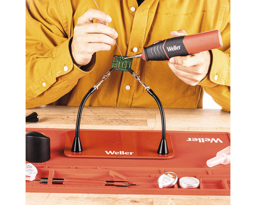 Szene zeigt das Löten einer Leiterplatte mit Lötzinn und Lötkolben, gehalten von einer Dritter-Hand-Löthilfe auf einer hitzebeständigen Arbeitsmatte von Weller.