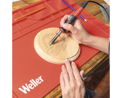 Eine Person verwendet einen Brennkolben von Weller auf einer Holzscheibe mit Anker-Motiv. Auf dem Tisch liegt eine rote Arbeitsmatte von Weller.