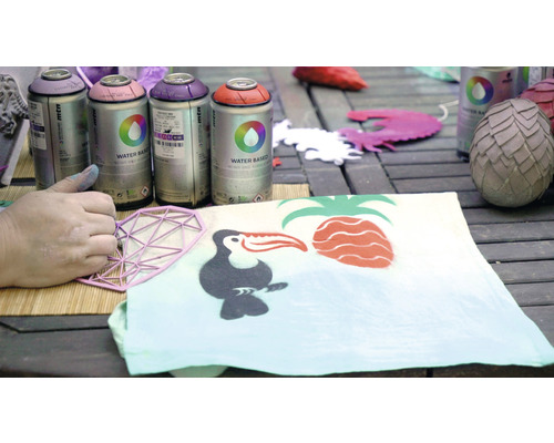 Sprühfarbe und Schablone beim Gestalten eines Stoffes mit tropischen Motiven