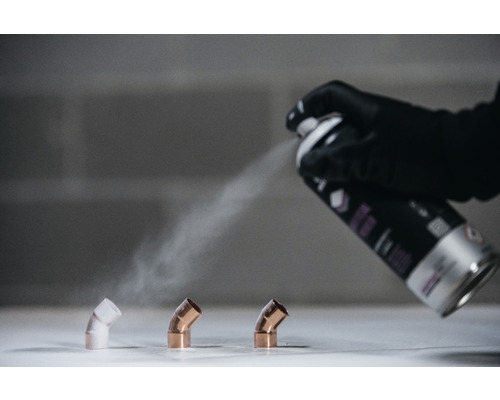 Eine Hand mit Handschuh sprüht Farbe auf Kupferbogenstücke mit einer Sprühdose.