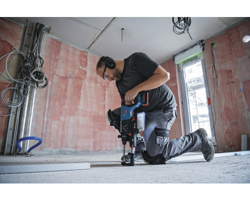 Ein Mann kniet und bohrt mit einem Bohrhammer in einen Betonboden in einem Raum im Rohbau.