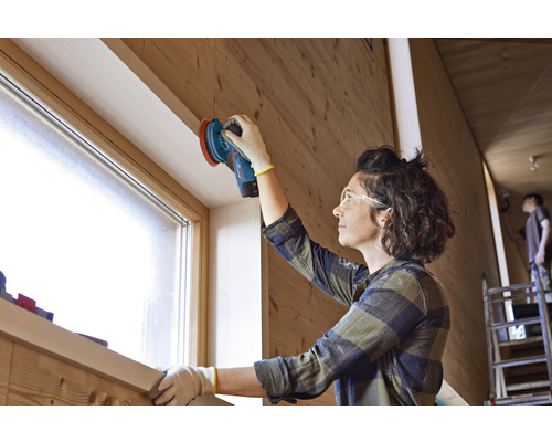 Frau schleift Holz mit einem Exzenterschleifer in einem hellen Raum.