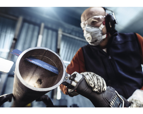 Mann mit Schutzbrille, Atemschutz, Gehörschutz und Handschuhen sägt ein Rohr mit einer Säbelsäge.