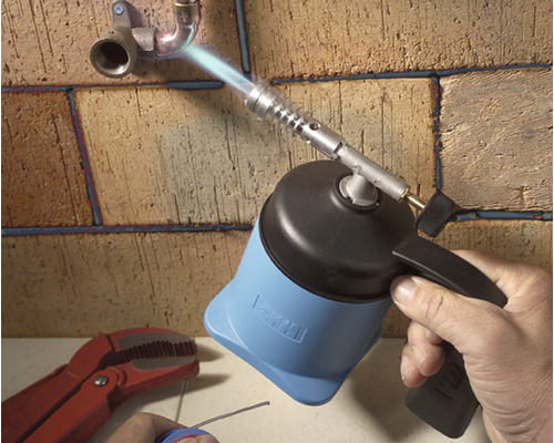 Eine Hand benutzt einen Brenner zum Löten von Rohren an einer Ziegelwand.