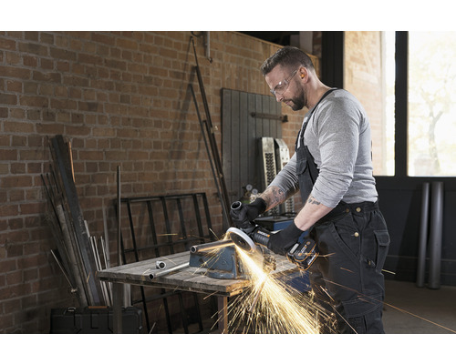 Ein Mann mit Schutzbrille bearbeitet ein Metallrohr mit einem Winkelschleifer in einer Werkstatt.