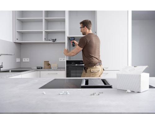 Ein Mann montiert eine Küche mit einem Akkuschrauber. Im Vordergrund sind Werkzeuge und Küchenutensilien zu sehen.