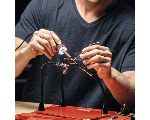 Person beim Löten mit einer Löthilfe und einem Lötkolben auf einer roten Arbeitsfläche