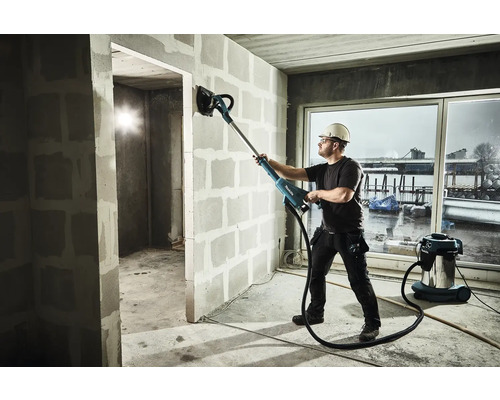 Ein Mann schleift eine Wand mit einem Langhalsschleifer in einem Rohbau, ein Staubsauger steht daneben.