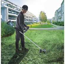 Ein Mann arbeitet mit einer Makita Akku-Sense auf einer großen Rasenfläche vor einem Wohnkomplex.