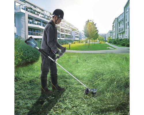 Ein Mann arbeitet mit einer Makita Akku-Sense auf einer großen Rasenfläche vor einem Wohnkomplex.