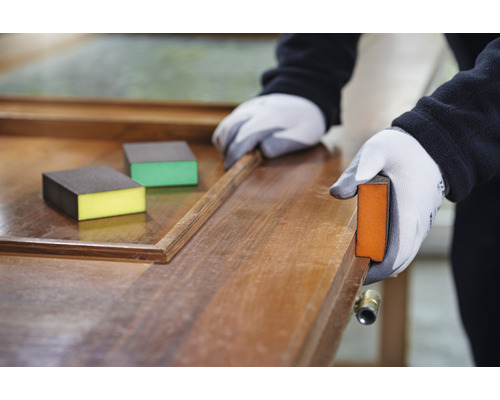 Eine Person schleift Holz mit einem Schleifklotz. Schleifschwämme liegen auf der Oberfläche.