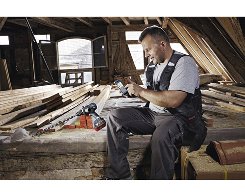 Ein Handwerker sitzt in einem Dachgeschoss und verwendet ein Smartphone, mit einem Akku-Bohrhammer der Marke Bosch neben sich.