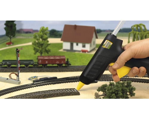 Modelleisenbahn-Szene mit Heißklebepistole in Aktion, umgeben von Miniaturhaus, Bäumen und Eisenbahnwaggons