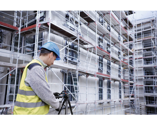 Bauarbeiter mit Helm und Warnweste bedient ein Laser-Distanzmessgerät auf einem Stativ vor einem Gebäude mit Gerüst.