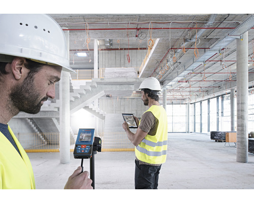 Zwei Bauarbeiter mit Helm und Sicherheitsweste nutzen einen Laser-Entfernungsmesser und ein Tablet auf einer Baustelle.
