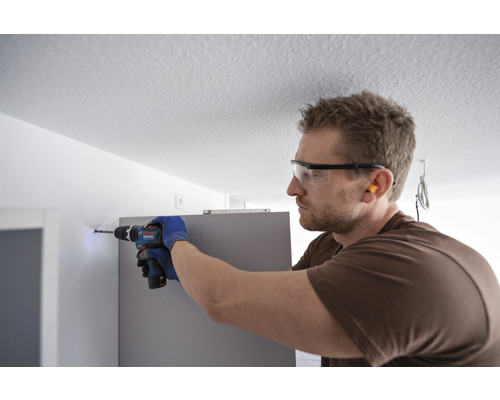 Ein Mann bohrt mit einer Bohrmaschine ein Loch in eine Wand neben einem Möbelstück. Er trägt eine Schutzbrille und Ohrstöpsel.