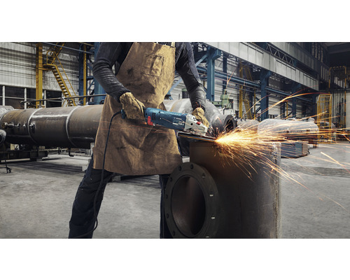 Ein Arbeiter in einer Werkshalle verwendet einen Winkelschleifer an einem Metallrohr, wobei Funken sprühen.