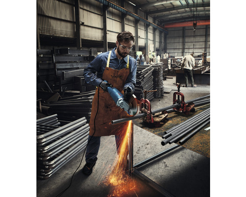 Ein Arbeiter in einer Fabrik schneidet mit einem Winkelschleifer ein Metallrohr, wobei Funken sprühen.