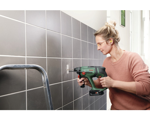 Eine Frau bohrt mit einer Bohrmaschine ein Loch in eine geflieste Wand. Sie trägt eine Schutzbrille und Ohrstöpsel.