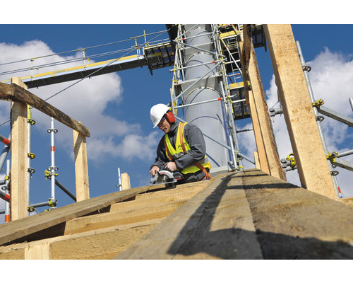 Ein Bauarbeiter mit Helm und Sicherheitsweste arbeitet mit einer Säge an einem Holzkonstruktionselement auf einer Baustelle mit Gerüst.