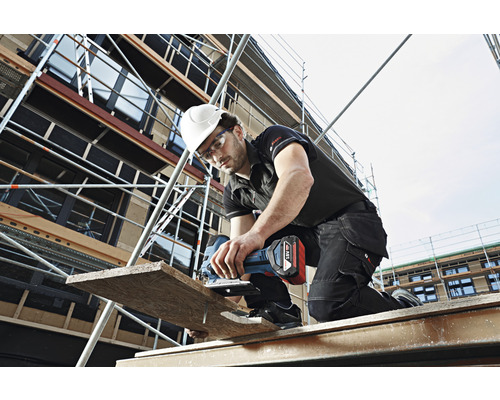 Handwerker mit Schutzhelm und Schutzbrille sägt ein Holzbrett mit einer Akku-Stichsäge auf einem Gerüst.