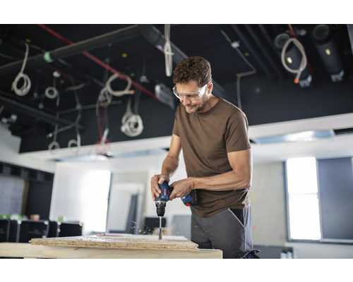 Ein Mann bohrt mit einer Bohrmaschine ein Loch in ein Holzbrett, während er eine Schutzbrille trägt.