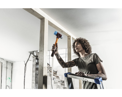 Eine Person steht auf einer Leiter und arbeitet mit einem Hammer an einem Holzrahmen im Innenbereich.