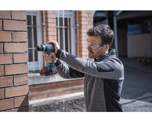 Mann bohrt mit Akku-Bohrschrauber in eine Ziegelwand, getragen werden Schutzbrille und Gehörschutzstöpsel