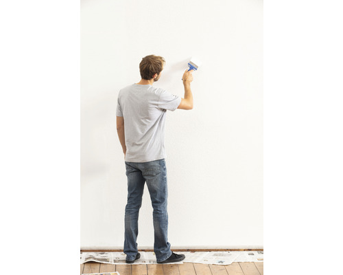 Mann streicht eine Wand mit einem Pinsel. Zeitungspapier schützt den Boden.