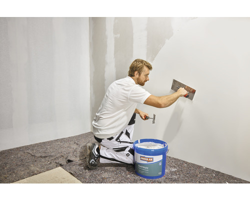 Mann trägt Glättputz mit einer Glättkelle auf eine Wand auf. Im Vordergrund steht ein Eimer Glättputz.