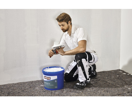 Mann trägt Mörtel mit einem Spachtel auf eine Wand auf. Im Vordergrund steht ein Eimer Mörtel.