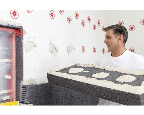 Ein Mann trägt eine Dämmplatte mit Kleber in einem Raum, der für Renovierungsarbeiten vorbereitet wird.