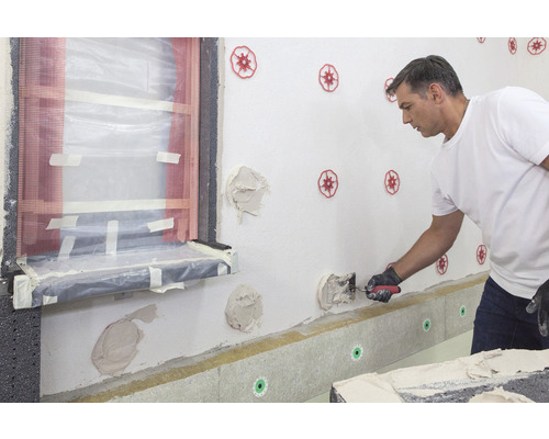 Ein Mann bringt Wärmedämmung an einer Hauswand an, wobei er eine Kelle und Dämmstoffdübel verwendet.