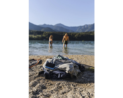 Kleiderhaufen und Lautsprecher am Strand mit Personen im Wasser und Bergen im Hintergrund