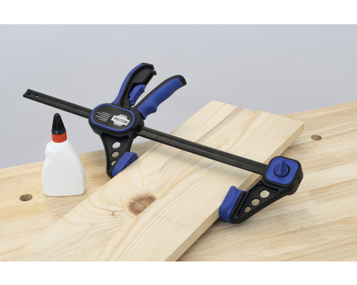 Eine INDUSTRIAL Einhandzwinge fixiert ein Holzbrett auf einer Werkbank, neben einer Flasche Holzleim.