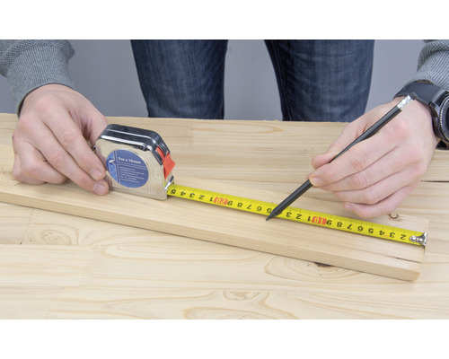 Eine Person misst ein Holzbrett mit einem Maßband und markiert es mit einem Bleistift.