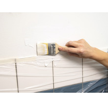 Eine Person streicht mit einem Pinsel eine Wand über Fliesen abgedeckt mit Folie.