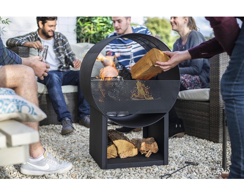 Gruppe von Leuten sitzt um eine Feuerstelle aus Metall mit Holzlagerung im Garten.