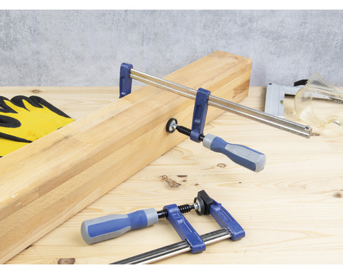 Holzstück mit Schraubzwingen, Arbeitshandschuhe, Tacker und Schutzbrille auf einem Holztisch