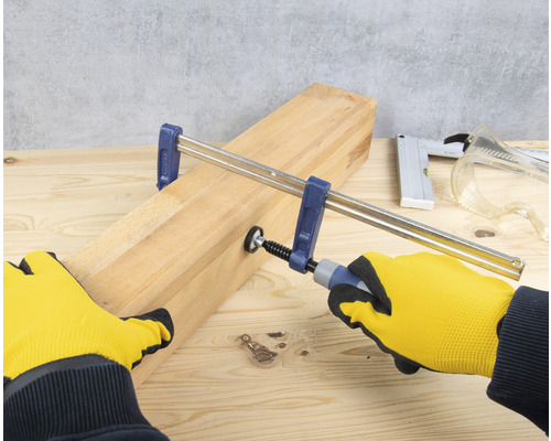 Eine Holzleiste wird mit einer Schraubzwinge fixiert. Werkzeuge liegen auf einer Holzoberfläche.