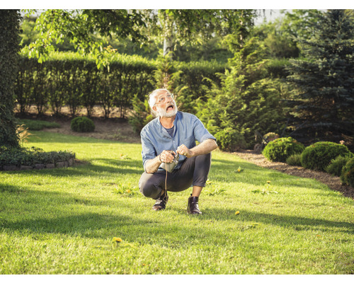 Ein Mann hockt auf einem Rasen mit einem Gartengerät in der Hand und blickt nach oben.