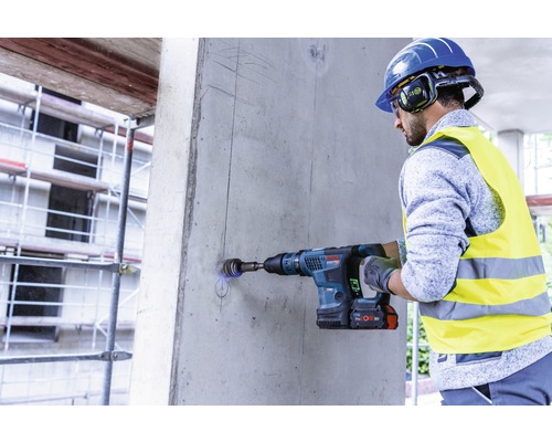 Ein Mann bohrt mit einem Akku Bohrhammer ein Loch in eine Betonwand. Der Mann trägt Sicherheitsausrüstung.