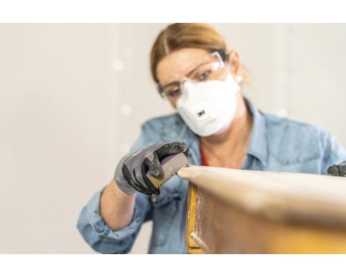 Frau schleift Holz mit Schleifpapier, Schutzbrille und Atemschutzmaske