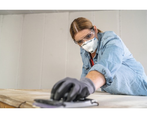 Frau schleift Holz mit Schleifklotz und trägt dabei Schutzbrille und Staubmaske