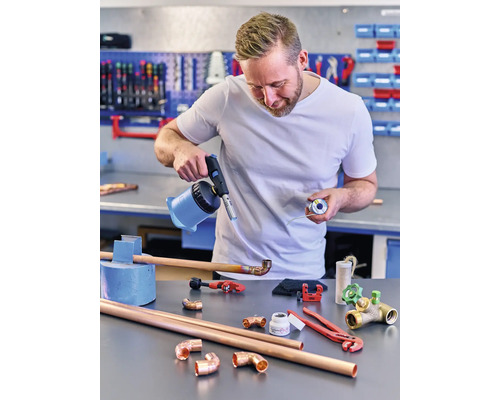 Mann lötet Kupferrohre mit einem Gasbrenner und Lötzinn auf einer Werkbank.