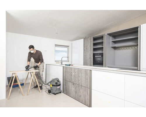 Ein Mann schleift Holz in einem Raum mit einer neuen Küche. Die Küche hat weiße und graue Elemente.