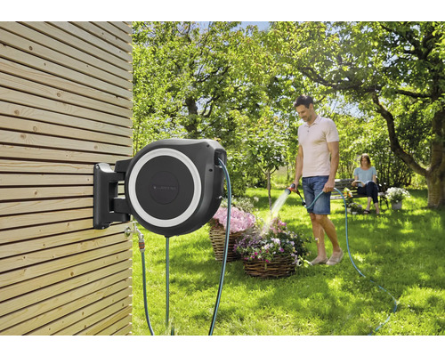 An einer Holzwand montierte Gartenschlauchbox mit Mann, der Blumen gießt.