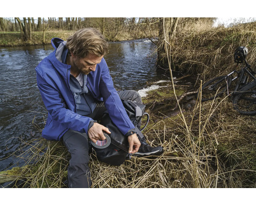 Mann mit Klebeband am Ufer eines Flusses, neben einem Fahrrad.