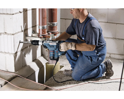 Ein Handwerker bohrt mit einem Bohrhammer ein Loch in eine Wand, Rohre verlaufen im Hintergrund.