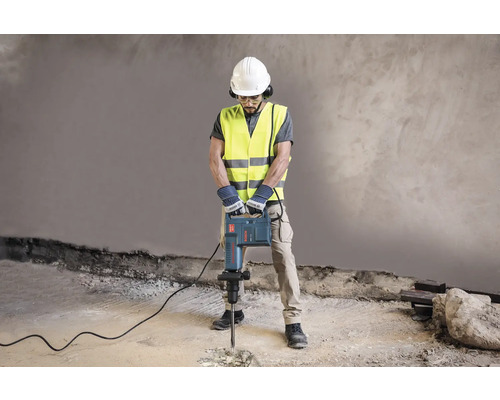Ein Bauarbeiter mit Schutzhelm, Schutzbrille, Gehörschutz, Schutzweste und Handschuhen bedient einen Presslufthammer.
